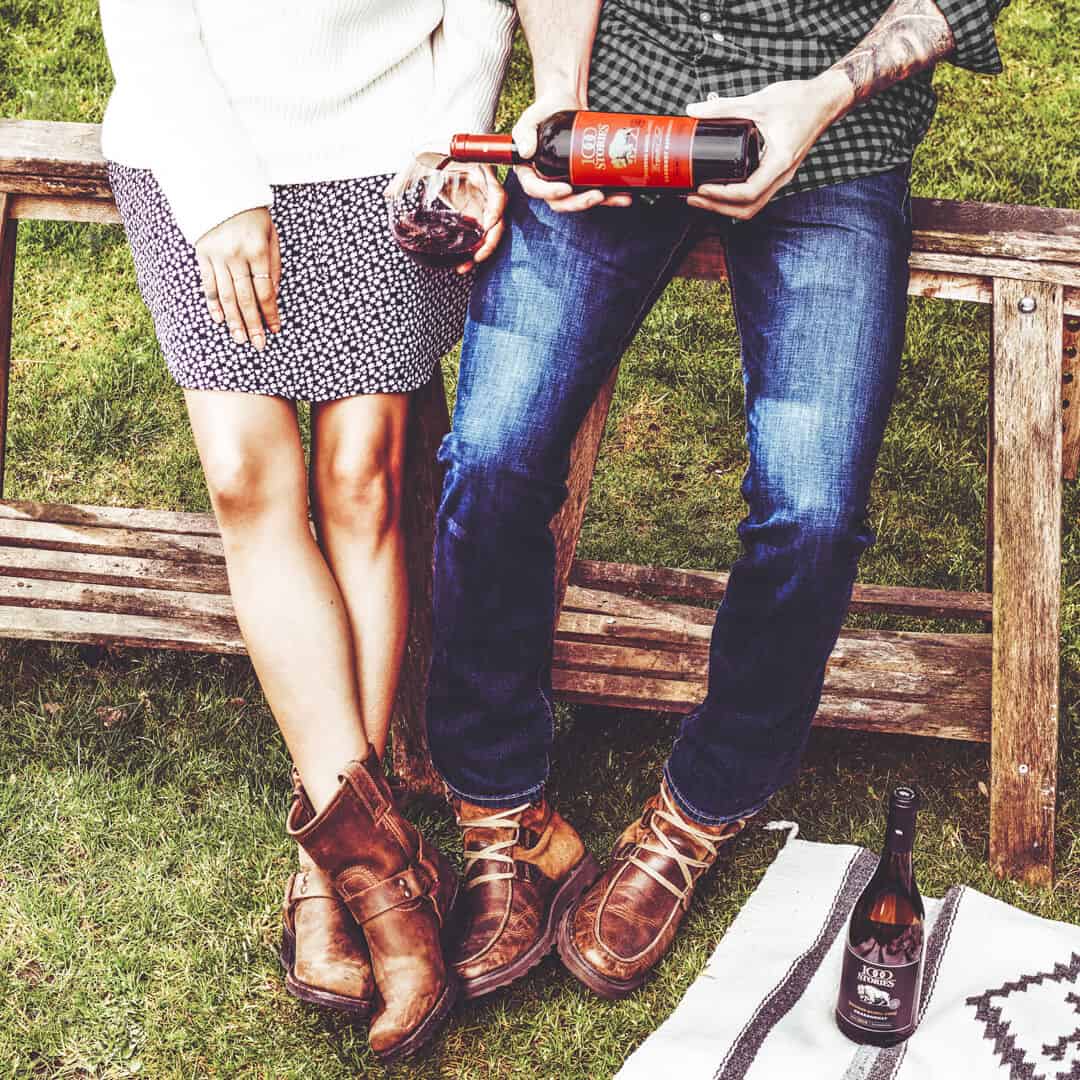 couple pours a glass of red wine while leaning on a wooden fence in a summery rural setting