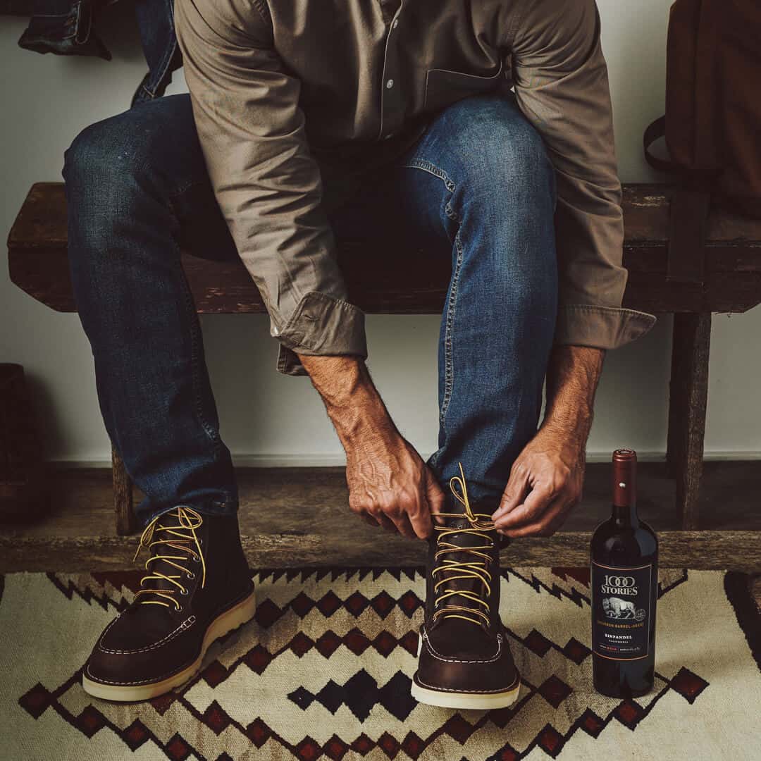 Man sitting on the bench tightening boot laces keeping 1000 Stories wine bottle next to his boot